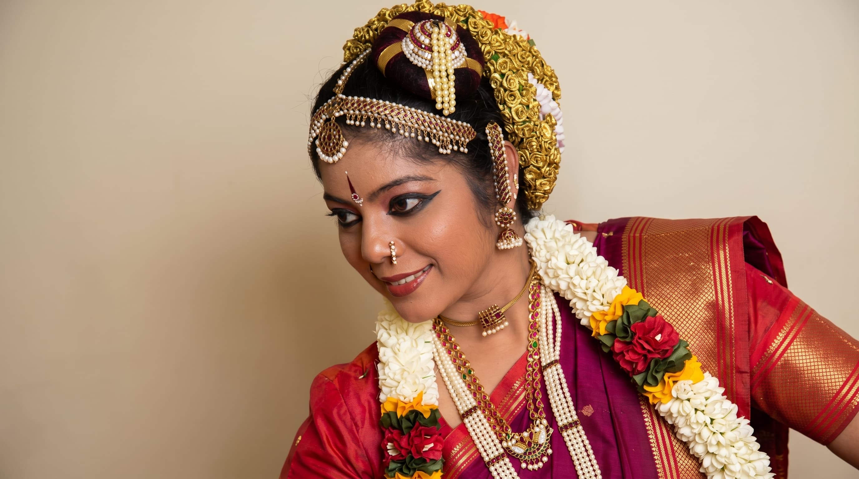 A Bharatanatyam dancer in bridal makeup and a gold temple headpiece woven with roses, jasmine and pearl strands draped over a magenta and red silk saree, smiling and looking down over her shoulder.